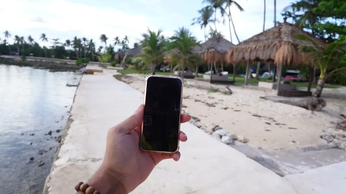 A hand holding a phone on the concrete path along the beach, with thatched huts ahead