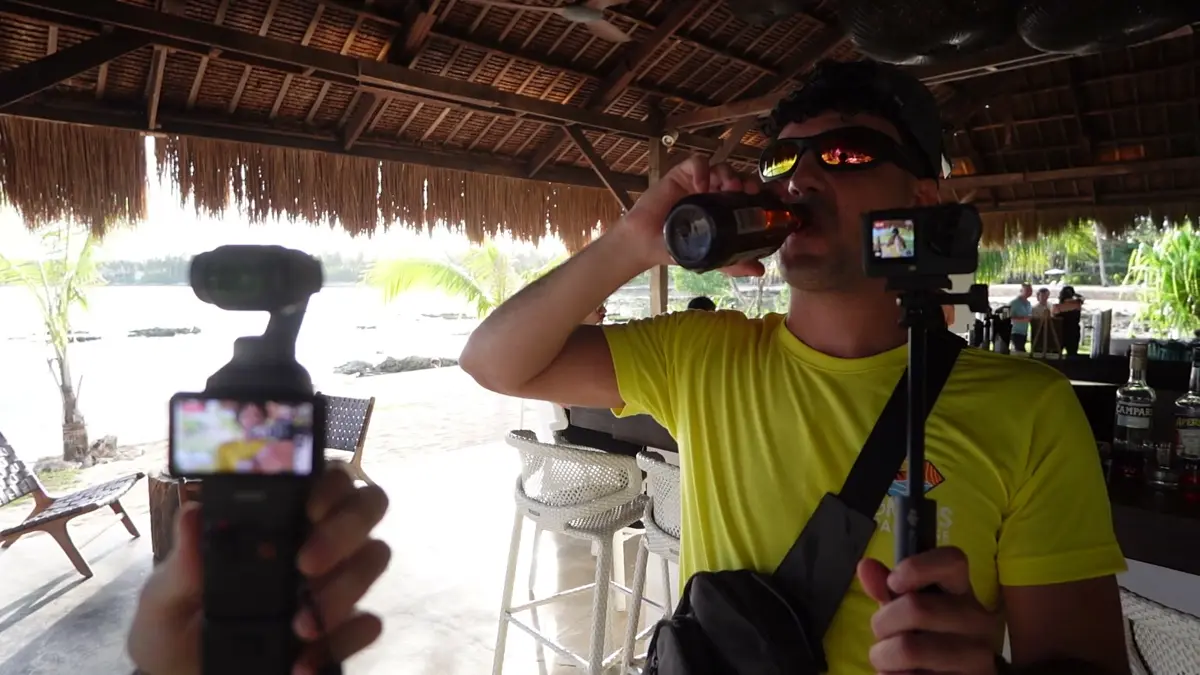 A Khun Camp crew member filming with a handheld camera under a thatched bar roof
